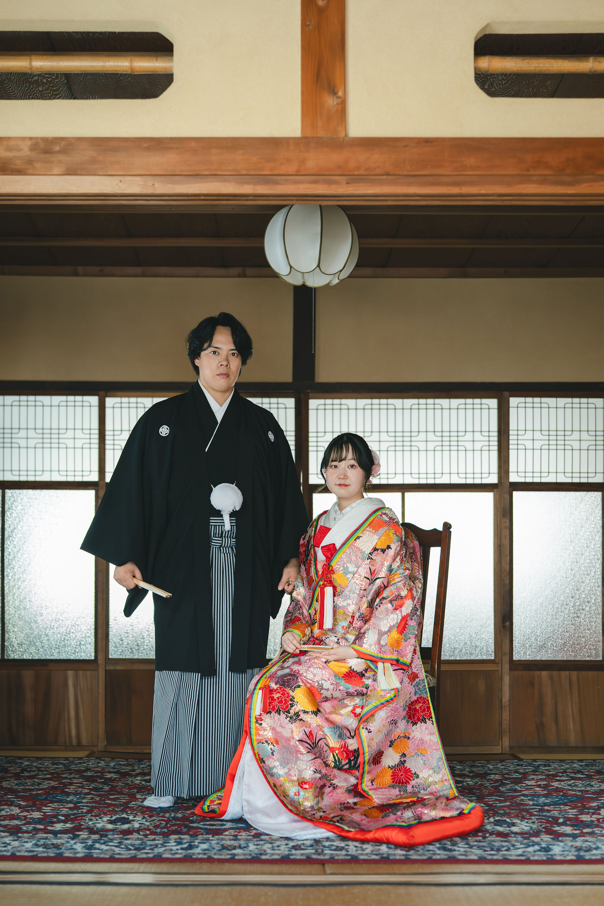 色打掛姿で座る花嫁と、その後ろに立つ黒紋付袴姿の新郎の和装前撮り写真