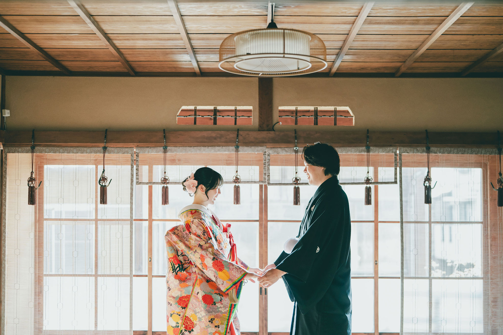 京町家の窓辺で、華やかな色打掛の花嫁と黒紋付袴の新郎が笑顔で向かい合い、手を取り合う和装前撮り写真。
